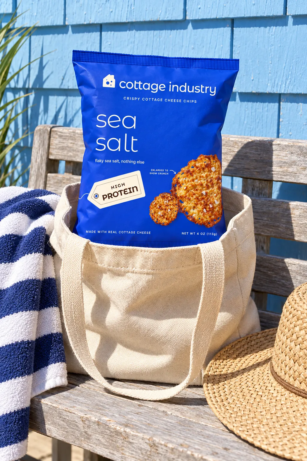 A bag of sea salt chips poking out of a canvas tote on a sun-bleached bench against a sky-blue shingle wall