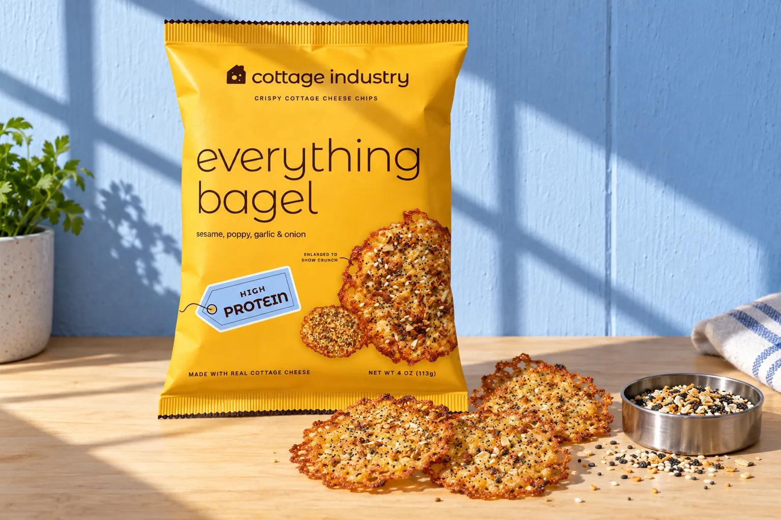 A bag of everything bagel chips on a pale maple counter against a powder-blue wall, three crisps and a tin of everything seasoning in front, a hard window shadow across the bag